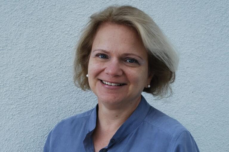 A smiling person with short blonde hair wearing a light blue collared shirt and pearl earrings stands against a textured light blue wall.