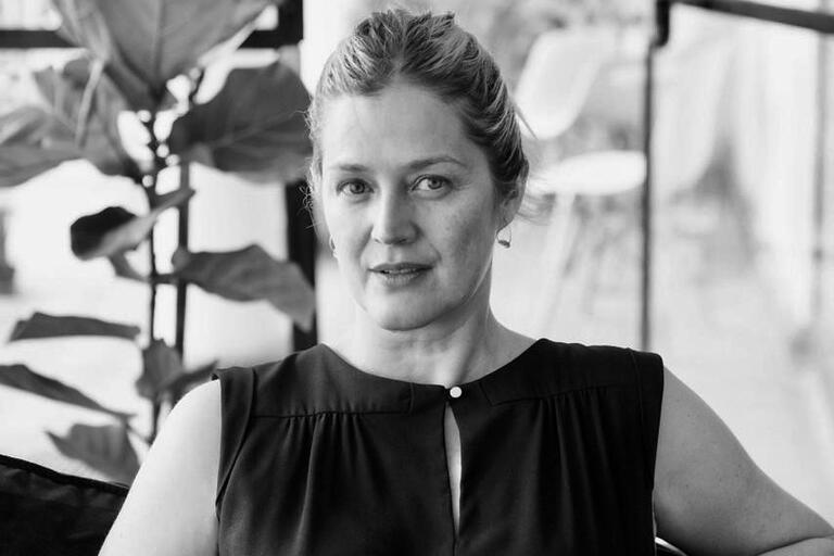 Black and white portrait of a woman with her hair pulled back, wearing a sleeveless top, seated indoors with plants and furniture in the background.
