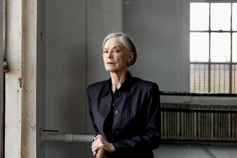 An older woman in a pinstriped suit stands indoors near a large window, holding onto a railing.