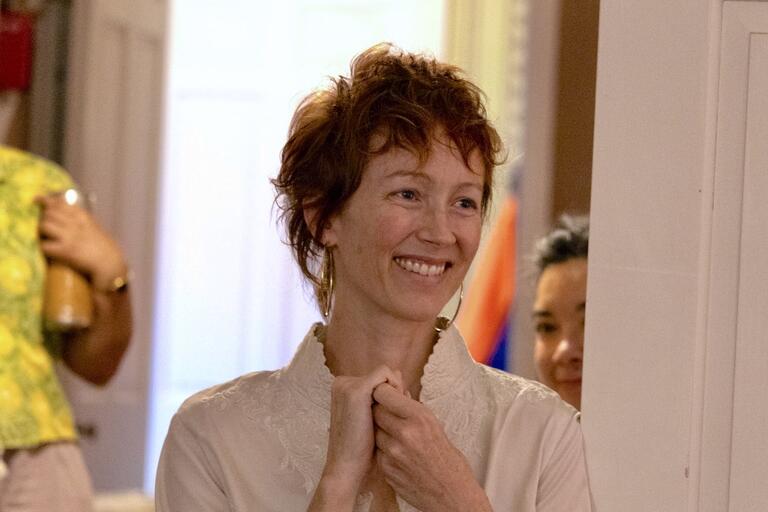 A person with short reddish-brown hair, wearing a light-colored patterned top, smiles while clasping their hands.