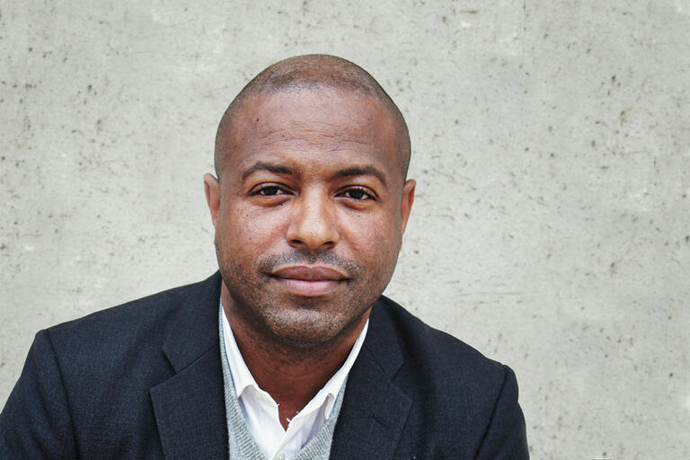 Portrait of a man against a grey textured wall, wearing a dark blazer, grey sweater, and white collared shirt.