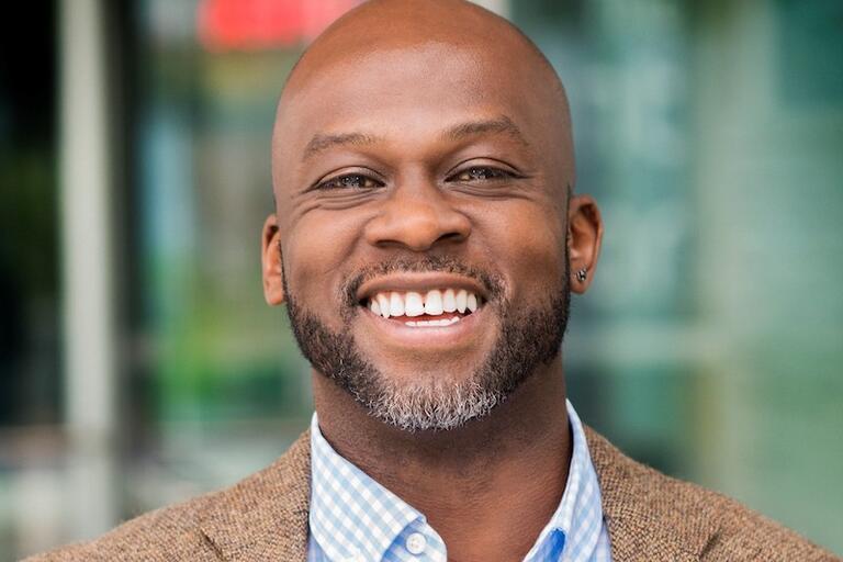 Close-up of a person smiling with a bald head, gray beard, and wearing a blue checkered shirt with a brown jacket.