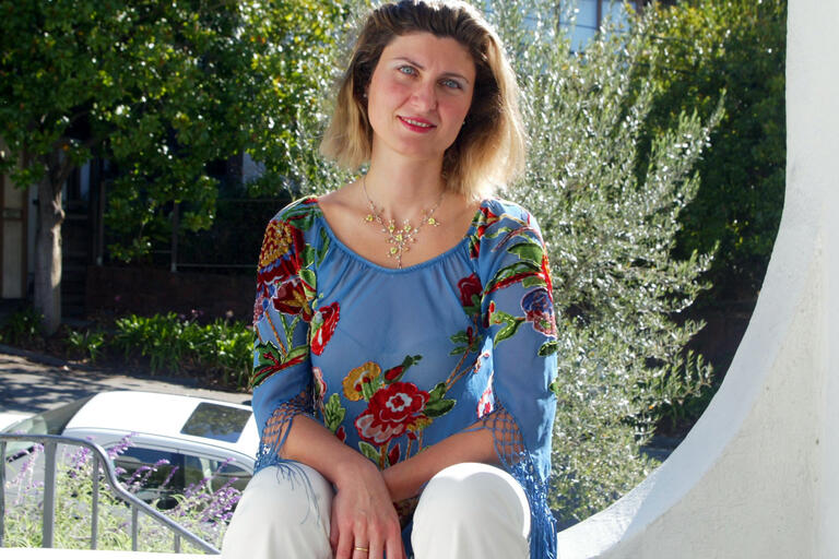 Woman sitting outdoors in a blue floral-embroidered top and white pants, with green trees and a white car in the background.