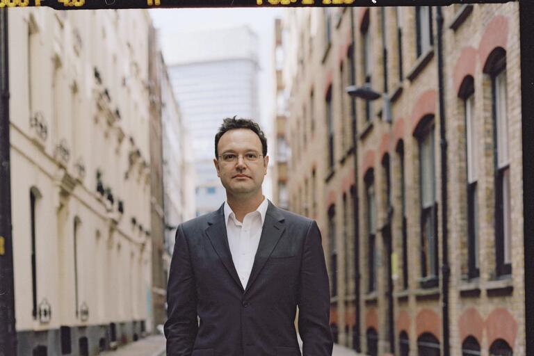 A man in a dark suit and white shirt stands in the middle of an alleyway lined with brick buildings, looking directly at the camera.