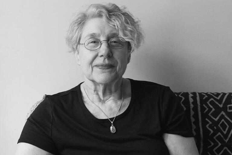 Black and white photo of an elderly woman wearing glasses and a dark shirt with a pendant necklace.