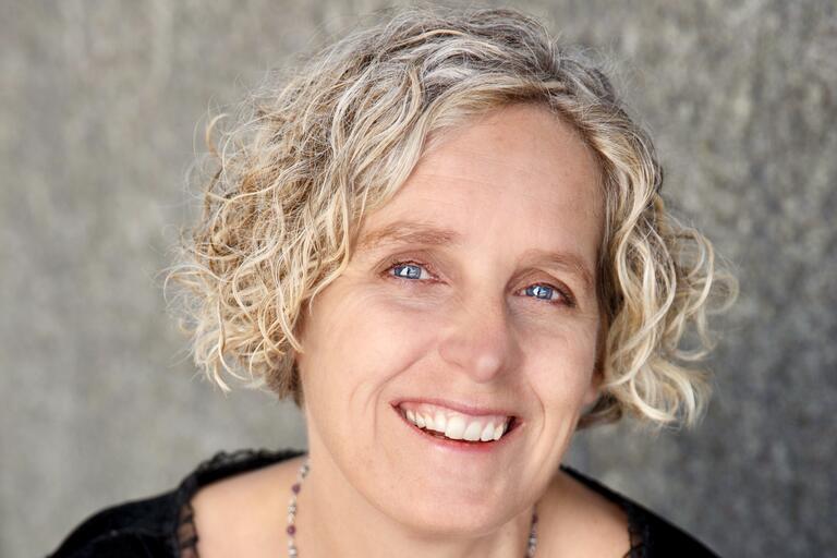Close-up of a smiling person with curly, blonde hair, blue eyes, and a black top, with a gray blurred background.