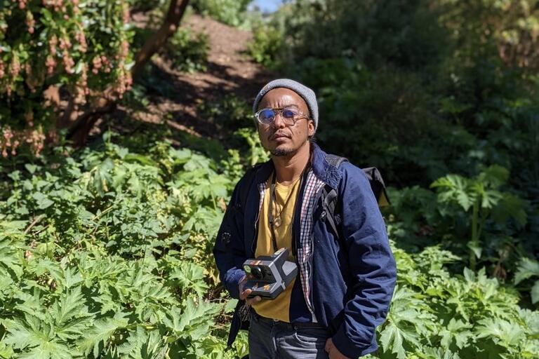Person in a blue jacket and gray beanie standing in a lush green garden holding a camera.