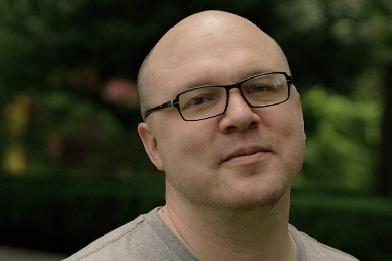 Close-up of a man with black glasses and a bald head, wearing a grey T-shirt, with a blurred green foliage background.