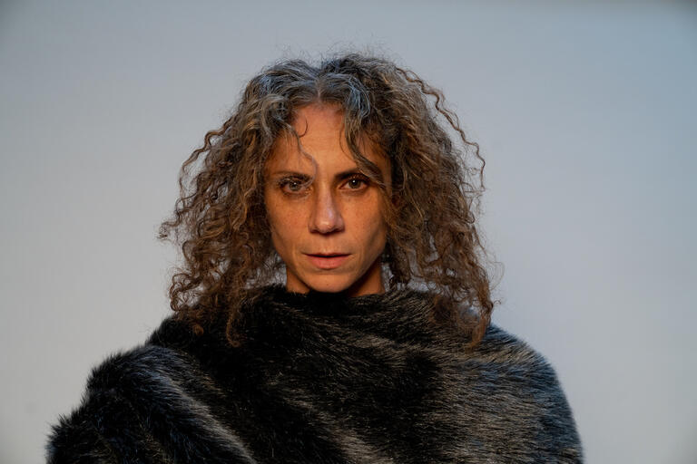 A person with curly hair wrapped in a dark, fur-like garment against a gray background.