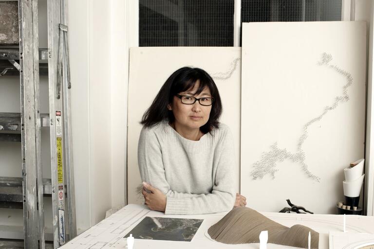 A woman in a gray sweater sits at a table with architectural models and drawings.
