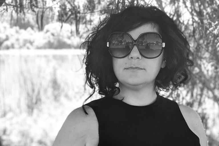 Black-and-white portrait of a person with curly hair and large sunglasses, wearing a sleeveless top in an outdoor setting with blurred trees in the background.