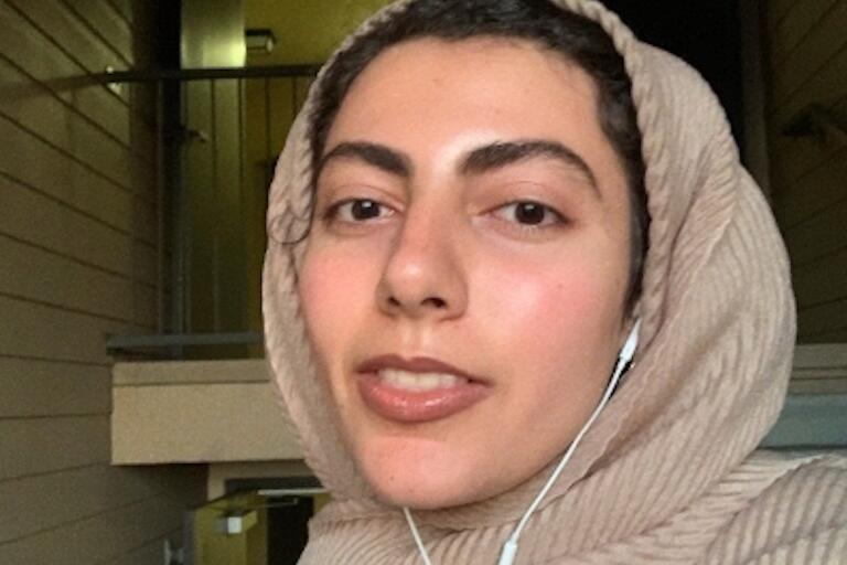 A woman in a tan headscarf and earbuds in looks at the camera