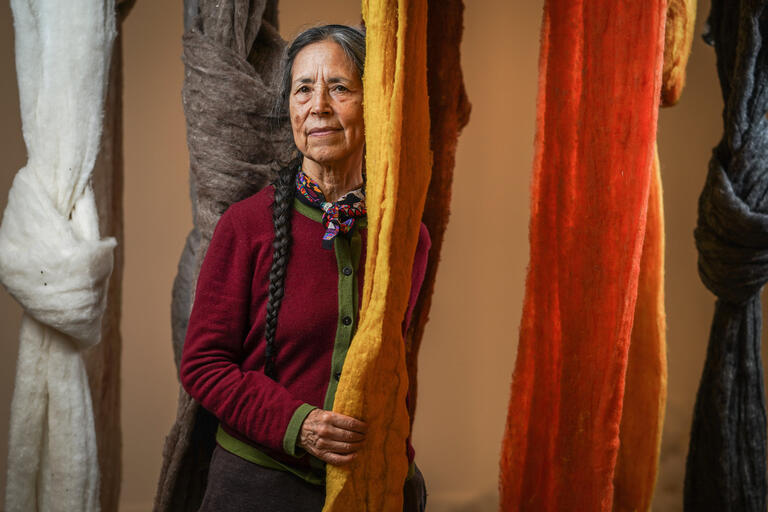 An older person stands among hanging textiles of various colors, including white, gray, yellow, orange, and black.
