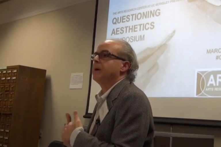 Man speaking at a podium with a presentation screen behind him displaying text about the "Questioning Aesthetics Symposium" at UC Berkeley.