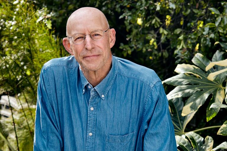 Portrait of an older person wearing a blue denim shirt, with a background of leafy plants and greenery.