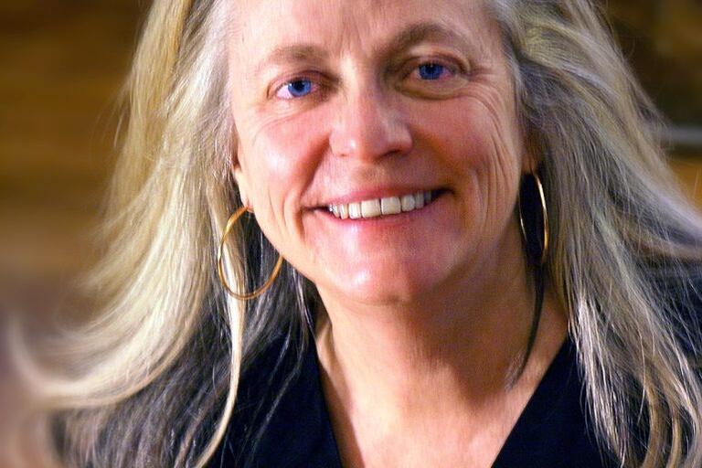 Middle-aged woman with long gray-blonde hair, wearing a dark V-neck top and gold hoop earrings, smiling toward the camera.