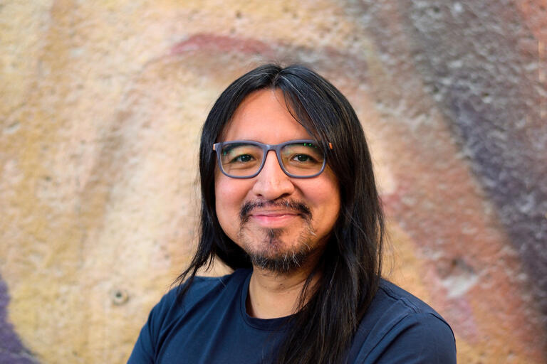 Person with long black hair and glasses smiling in front of a textured, earthy-toned wall.