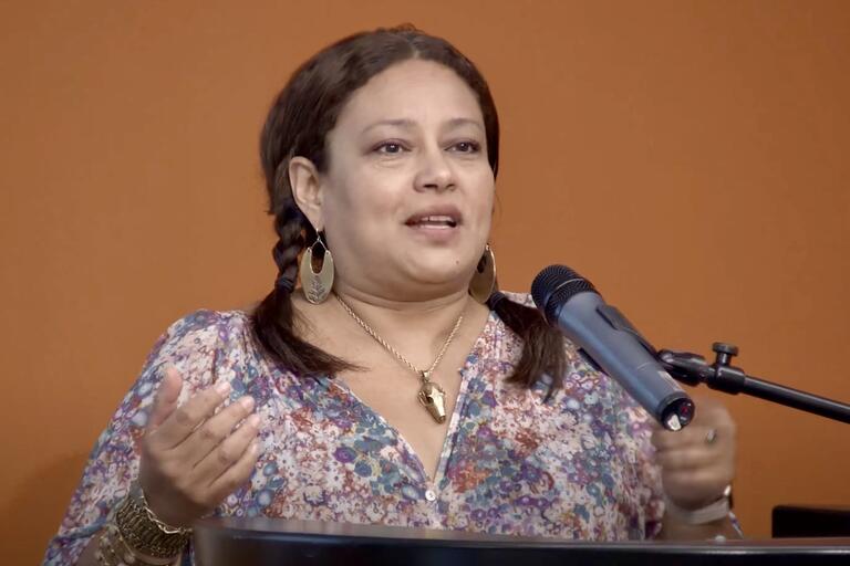 Person speaking at a podium with a microphone, wearing a floral blouse, large earrings, and a necklace.