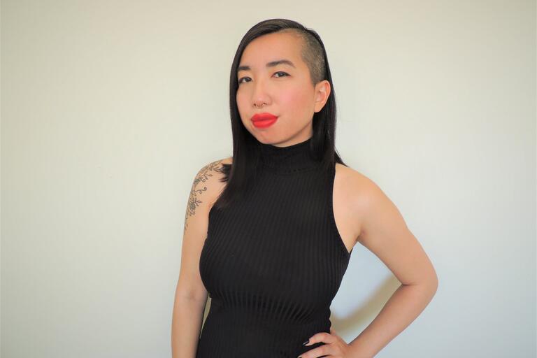 Person with medium-length black hair shaved on one side, wearing a black sleeveless dress, red lipstick, and septum piercing, standing against a white background.