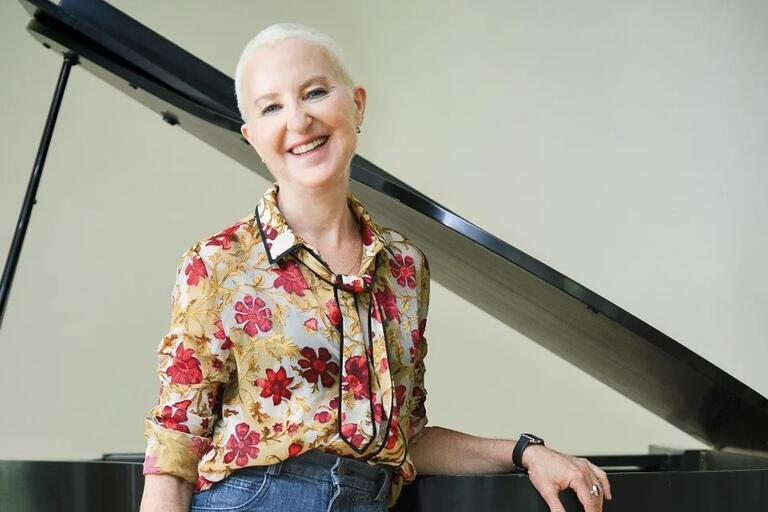 A person in a floral blouse and jeans standing beside a grand piano.