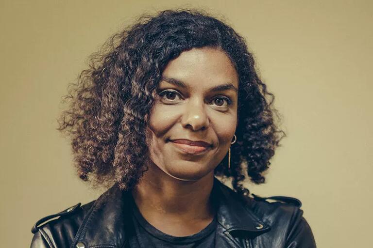 Portrait of a person with curly hair wearing a black leather jacket and a dark shirt against a beige background.