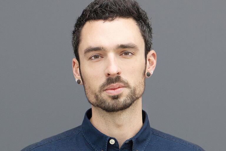 Portrait of an individual with short curly hair, a beard, and wearing a navy blue buttoned shirt with gauge earrings. Plain gray background.