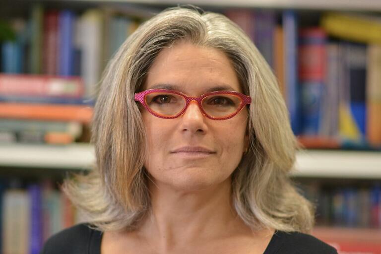Person with grey hair and red-rimmed glasses in front of bookshelves.