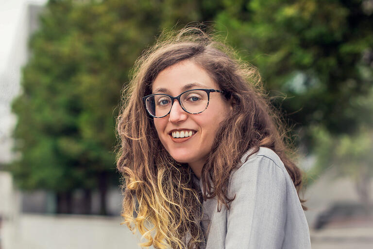 Person with long, curly brown to blonde hair, wearing glasses and a light gray jacket, smiling against a background of green trees and a blurred building.