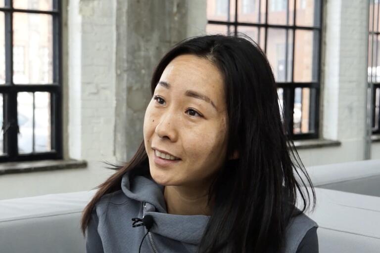 Close-up of a woman with long black hair in a gray hoodie, with large industrial windows in the background.
