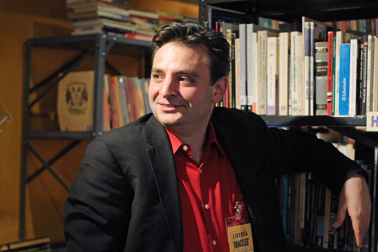 A man in a dark blazer and red shirt seated in front of bookshelves filled with books.