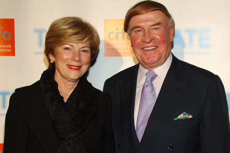 A man in a suit and a woman in a black outfit smiling in front of a backdrop featuring the "cifo" and "TATE" logos.