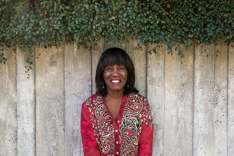 Woman standing in front of a wooden fence with greenery above, wearing a red patterned blouse and smiling.
