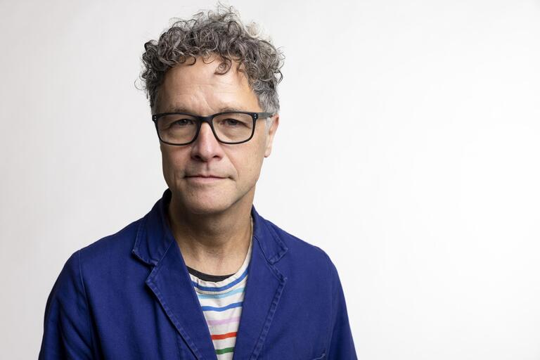 Middle-aged man with curly, graying hair, wearing black-framed glasses, a blue jacket, and a striped shirt, standing against a plain light background.
