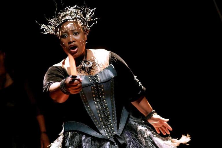 Performer on stage holding a prop knife, wearing a black costume with a decorative bodice and headpiece, under dramatic lighting.