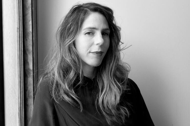 Black-and-white portrait of a person with long wavy hair, near a wooden-framed window.
