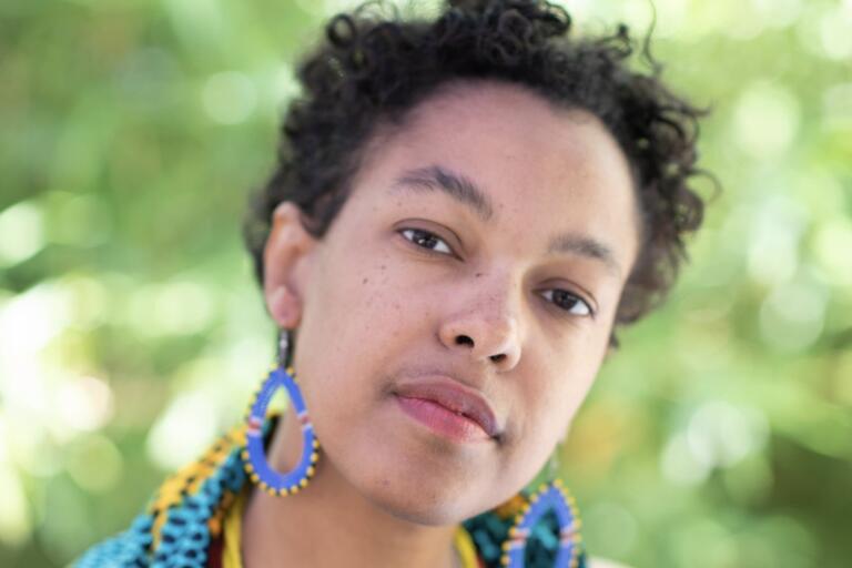 Close-up portrait of a person with short curly hair, wearing colorful teardrop-shaped earrings against a blurred green foliage background.