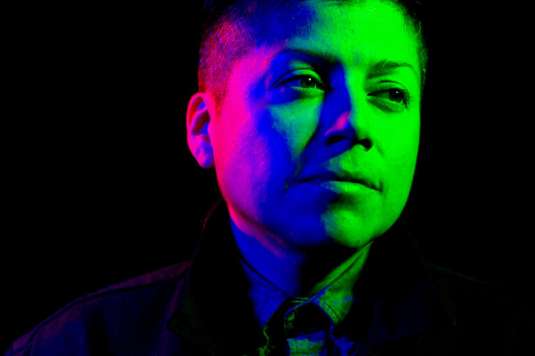 Close-up portrait of a person with short hair, illuminated with green and pink lighting against a black background.