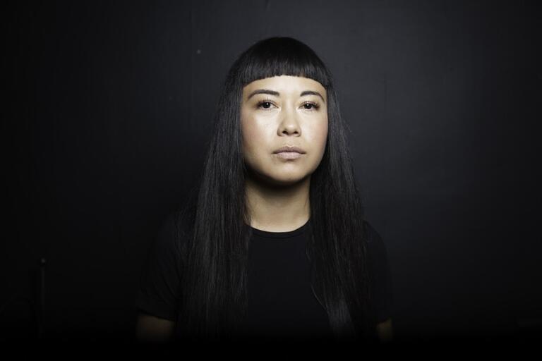 Portrait of a person with long black hair and blunt bangs, wearing a black shirt, against a black background.
