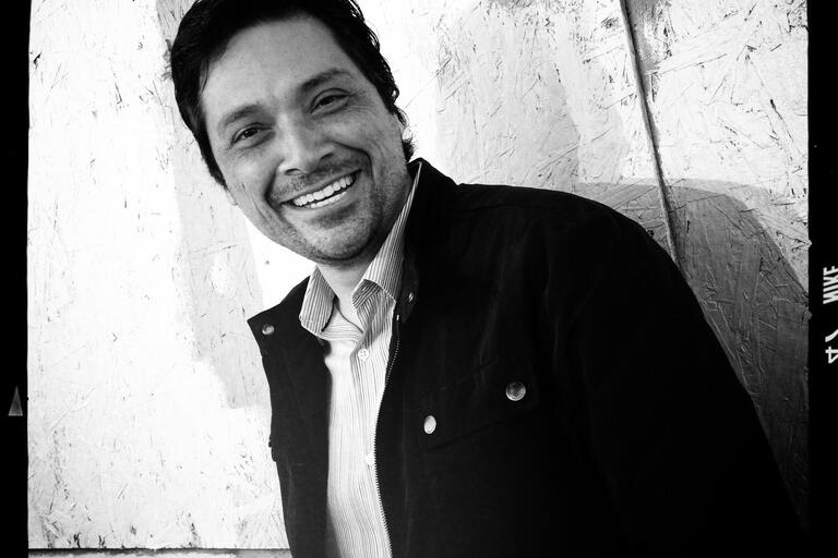 Black-and-white photo of a man smiling and leaning against a textured wall background.