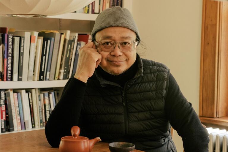 Person with a gray beanie and glasses, smiling and resting their cheek on their hand, in front of a bookshelf, with a teapot and bowl on the table.
