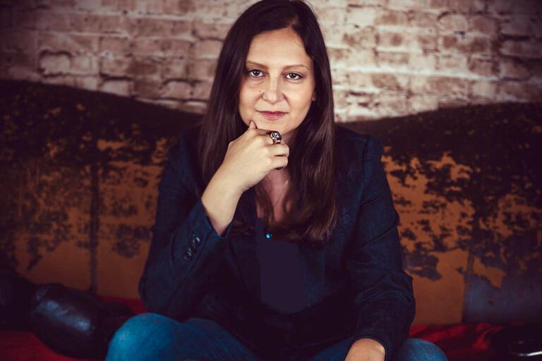 A woman with long brown hair in a dark blazer and blue jeans sits in front of a textured brick wall.