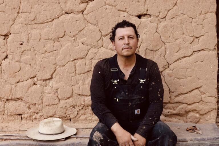 A man in black overalls sits in front of a cracked adobe wall with a white hat beside him.