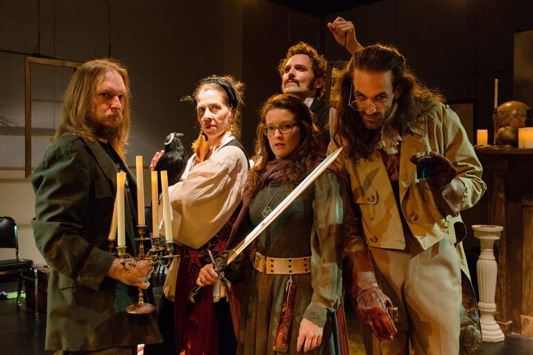 ive people in theatrical costumes, with props including a candelabrum, black bird, sword, and glass jar.