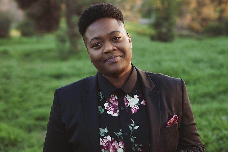 Person standing in a grassy field wearing a black blazer and a floral patterned shirt.