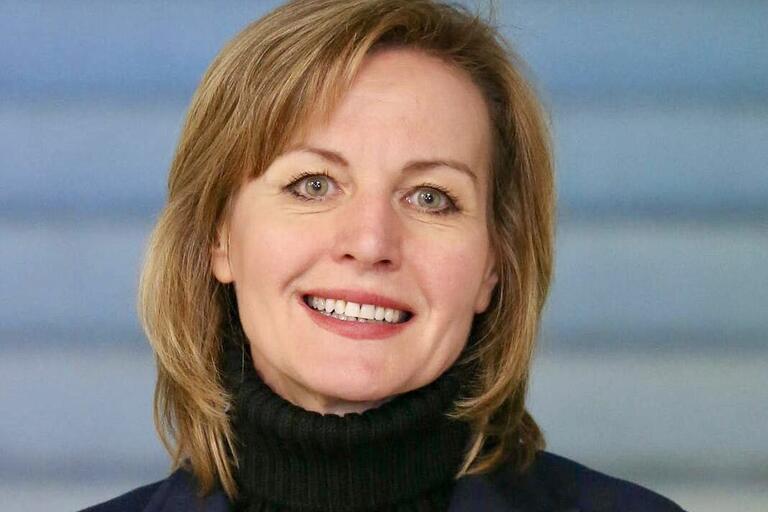 Portrait of a smiling woman with shoulder-length light brown hair, wearing a black turtleneck and dark blazer, against a blue-toned background.