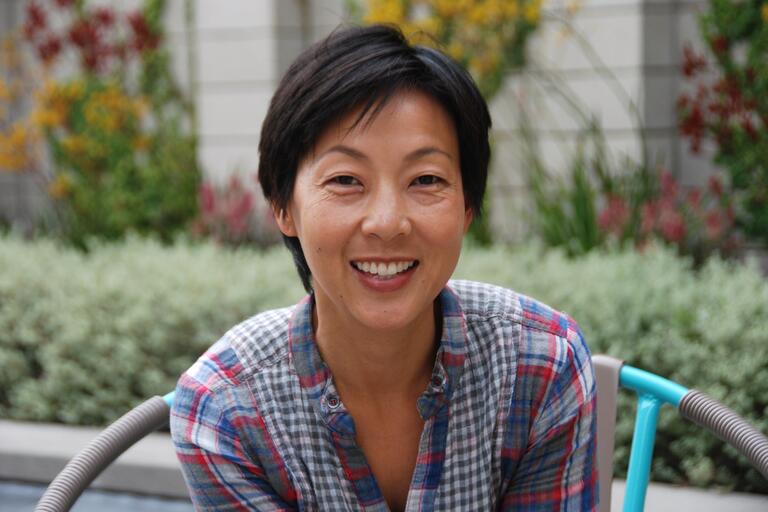 Person with short dark hair smiling, wearing a plaid shirt, sitting outdoors with a garden in the background.