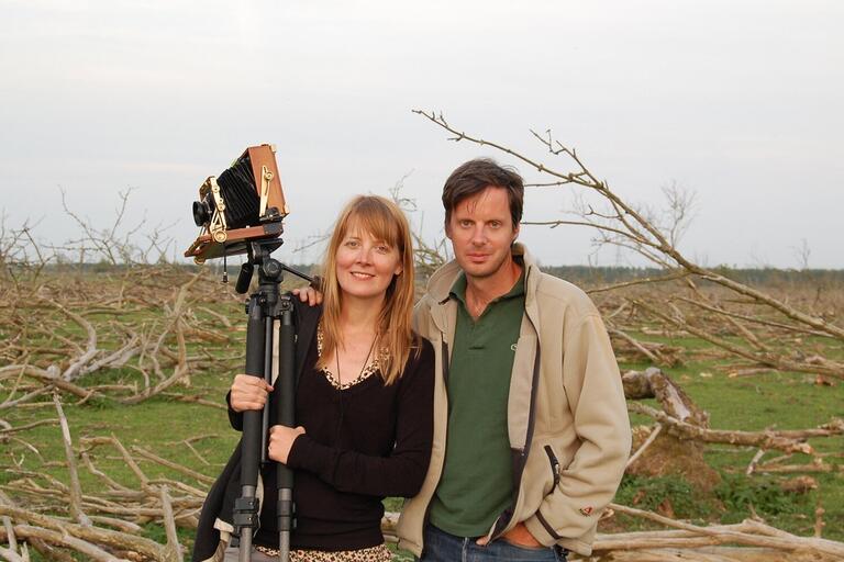 Two people stand in a field of fallen branches, holding a large-format camera on a tripod.