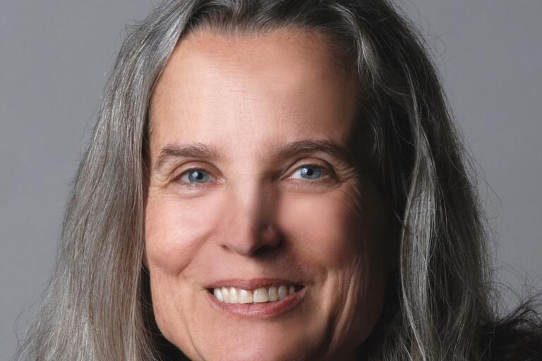 Close-up of a smiling person with long gray hair and light blue eyes against a gray background.