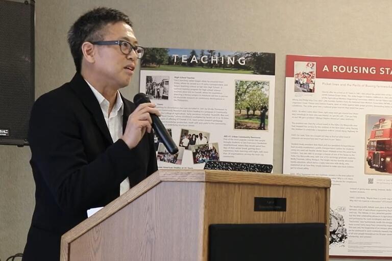 A person in a black blazer speaks into a microphone behind a wooden podium. There are informational posters on the wall behind him.