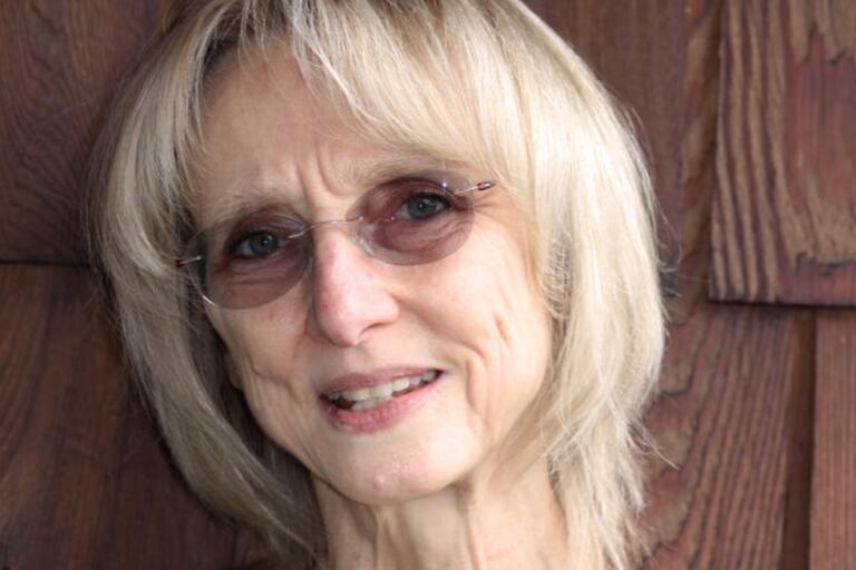 A woman in a black sweater with blond hair and glasses looks into the camera.